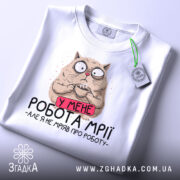 Футболка робота мрії, білого кольору, складена на столі, фон світло-сірий, принт з котом, який тримає чашку, з текстом 'У МЕНЕ РОБОТА МРІЇ - АЛЕ Я НЕ МРІЯВ ПРО РОБОТУ', з етикеткою зеленого кольору.