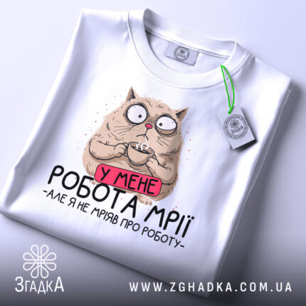 Футболка робота мрії, білого кольору, складена на столі, фон світло-сірий, принт з котом, який тримає чашку, з текстом 'У МЕНЕ РОБОТА МРІЇ - АЛЕ Я НЕ МРІЯВ ПРО РОБОТУ', з етикеткою зеленого кольору.