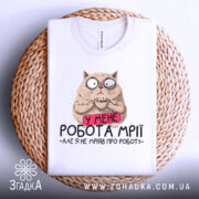 Футболка робота мрії, білого кольору, складена на плетеній кошику, фон світло-сірий, принт з котом, який тримає чашку, з текстом 'У МЕНЕ РОБОТА МРІЇ - АЛЕ Я НЕ МРІЯВ ПРО РОБОТУ'.