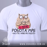 Футболка робота мрії, білого кольору, складена на столі, фон світло-сірий, принт з котом, який тримає чашку, з текстом 'У МЕНЕ РОБОТА МРІЇ - АЛЕ Я НЕ МРІЯВ ПРО РОБОТУ', з етикеткою зеленого кольору.