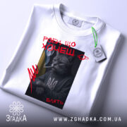 Футболка роби що хочеш, біла, складена на поверхні, фон світло-сірий, принт з котом у свитері з тризубом, котяча фігура з сигаретою, текст червоним кольором, на комірці видна етикетка.