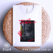 Футболка роби що хочеш, біла, складена на плетеній кошику, фон світло-сірий, принт з котом у свитері з тризубом, котяча фігура з сигаретою, текст червоним кольором.