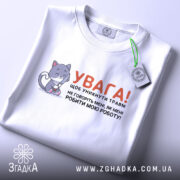 Футболка робити мою роботу. Біла футболка, складена на поверхні, з видимим коміром. На фоні світло-сірий колір. Принт з малюнком котика та текстом, що закликає до обережності, прикріплений тегом.