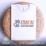 Футболка робити мою роботу. Біла футболка, складена на плетеній кошику, з видимим коміром. На фоні світло-сірий колір. Принт містить малюнок котика та текст, що закликає до обережності.