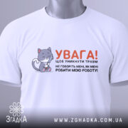 Футболка робити мою роботу. Біла футболка, складена на поверхні, з видимим коміром. На фоні світло-сірий колір. Принт з малюнком котика та текстом, що закликає до обережності, прикріплений тегом.