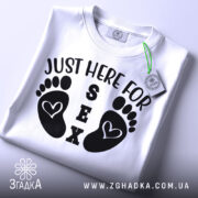 Футболка родинний гумор, біла, складена на столі, фон світлий, принт з чорними слідами ніг і текстом 'JUST HERE FOR SEX', на комірці видна етикетка.