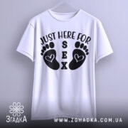 Футболка родинний гумор, біла, підвішена на вішалці, фон світло-сірий, принт з чорними слідами ніг і текстом 'JUST HERE FOR SEX'.