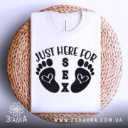 Футболка родинний гумор, біла, складена на плетеній кошику, фон світлий, принт з чорними слідами ніг і текстом 'JUST HERE FOR SEX'.