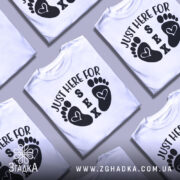 Футболка родинний гумор, біла, зображена на манекені, фон нейтральний, принт з чорними слідами ніг і текстом 'JUST HERE FOR SEX'.