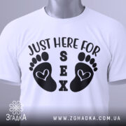 Футболка родинний гумор, біла, складена на столі, фон світлий, принт з чорними слідами ніг і текстом 'JUST HERE FOR SEX', на комірці видна етикетка.