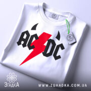 Футболка рок енергія, біла, складена на поверхні, фон світло-сірий, принт з червоним блискавкою та чорними літерами 'AC/DC', з етикеткою на зеленому шнурку.
