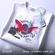 Футболка рок естетика, біла, складена на поверхні, злегка зверху, фон світло-сірий, принт з черепом, червоними крилами та текстом, прикріплена етикетка з зеленим шнурком.