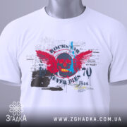 Футболка рок естетика, біла, складена на поверхні, злегка зверху, фон світло-сірий, принт з черепом, червоними крилами та текстом, прикріплена етикетка з зеленим шнурком.