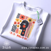 Футболка розквіт особистості. Біла футболка, складена на столі, з етикеткою, що звисає. На фоні світло-сірий колір. Принт з абстрактним обличчям, оточеним кольоровими квітами, виконаний в яскравих кольорах.