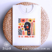 Футболка розквіт особистості. Біла футболка, складена на плетеному кошику. На фоні світло-сірий колір. Принт містить абстрактне обличчя з чорними рисами та яскравими квітами, виконаний в оранжевих і жовтих відтінках.