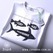 Футболка розмір має значення, біла, складена на поверхні, видно етикетку з зеленим шнурком, принт з рибами та написами "РОЗМІР", "МАС" і "ЗНАЧЕННЯ" у чорному кольорі, фон світлий.