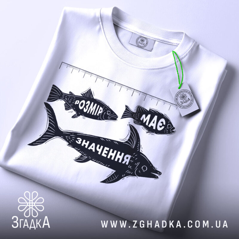 Футболка розмір має значення, біла, складена на поверхні, видно етикетку з зеленим шнурком, принт з рибами та написами "РОЗМІР", "МАС" і "ЗНАЧЕННЯ" у чорному кольорі, фон світлий.