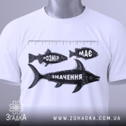 Футболка розмір має значення, біла, складена на поверхні, видно етикетку з зеленим шнурком, принт з рибами та написами "РОЗМІР", "МАС" і "ЗНАЧЕННЯ" у чорному кольорі, фон світлий.