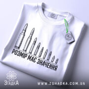 Футболка розмір має значення, біла, складена на столі, фон світлий, принт з дев'яти патронів, текст чорного кольору, на футболці є етикетка з зеленим шнурком.