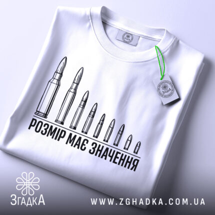 Футболка розмір має значення, біла, складена на столі, фон світлий, принт з дев'яти патронів, текст чорного кольору, на футболці є етикетка з зеленим шнурком.