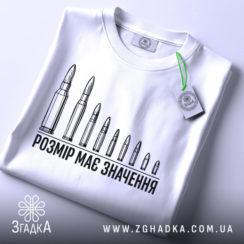 Футболка розмір має значення, біла, складена на столі, фон світлий, принт з дев'яти патронів, текст чорного кольору, на футболці є етикетка з зеленим шнурком.