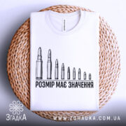 Футболка розмір має значення, біла, складена на плетеній кошику, фон світлий, принт з дев'яти патронів, текст чорного кольору, видно етикетку на горловині.