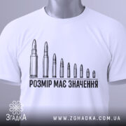 Футболка розмір має значення, біла, складена на столі, фон світлий, принт з дев'яти патронів, текст чорного кольору, на футболці є етикетка з зеленим шнурком.