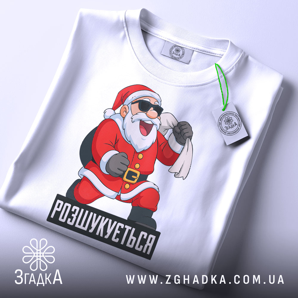 Футболка розшукується Санта. Біла футболка, зображена зверху, з принтом Санти, який сміється, тримаючи мішок. На футболці є етикетка з зеленим шнурком. Фон світлий, однорідний.