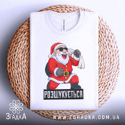 Футболка розшукується Санта. Біла футболка, складена на плетеній основі, з принтом Санти, який сміється. Фон світлий, з текстурою плетеного матеріалу.