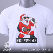 Футболка розшукується Санта. Біла футболка, зображена зверху, з принтом Санти, який сміється, тримаючи мішок. На футболці є етикетка з зеленим шнурком. Фон світлий, однорідний.