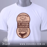 Футболка рука допомоги, білого кольору, розкладена на поверхні, фон світло-сірий, принт коричневого кольору з текстом та зображенням руки і ноги, на футболці є етикетка з зеленим шнурком.