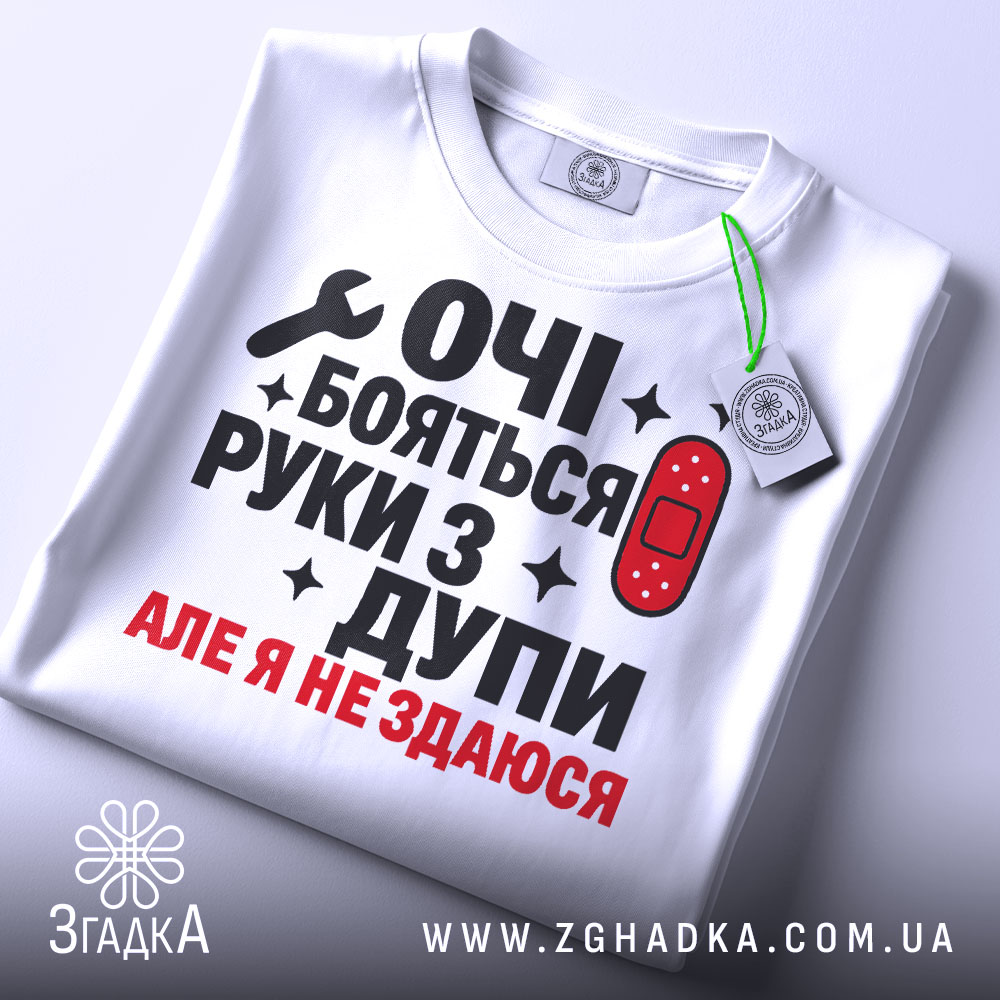 Футболка руки з дупи, біла, складена на столі, фон світло-сірий, принт чорного та червоного кольору з текстом "ОЧІ БОЯТЬСЯ РУКИ З ДУПИ АЛЕ Я НЕ ЗДАЮСЯ" та зображенням ключа і пластиру, на комірці є етикетка.