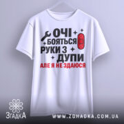 Футболка руки з дупи, біла, підвішена на вішалці, фон світло-сірий, принт чорного та червоного кольору з текстом "ОЧІ БОЯТЬСЯ РУКИ З ДУПИ АЛЕ Я НЕ ЗДАЮСЯ" та зображенням ключа і пластиру.