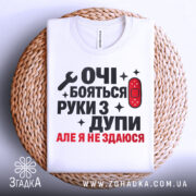 Футболка руки з дупи, біла, складена на плетеній кошику, фон світлий, принт чорного та червоного кольору з текстом "ОЧІ БОЯТЬСЯ РУКИ З ДУПИ АЛЕ Я НЕ ЗДАЮСЯ" та зображенням ключа і пластиру.