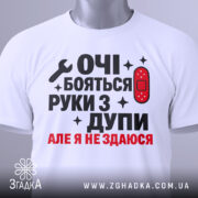Футболка руки з дупи, біла, складена на столі, фон світло-сірий, принт чорного та червоного кольору з текстом "ОЧІ БОЯТЬСЯ РУКИ З ДУПИ АЛЕ Я НЕ ЗДАЮСЯ" та зображенням ключа і пластиру, на комірці є етикетка.