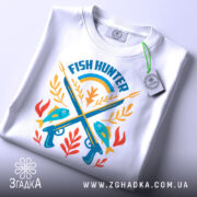 Футболка рибалка мисливець, біла, зображена зверху, фон світло-сірий, принт з рибами, гарпунами та рослинами, прикріплена етикетка з зеленим шнурком.