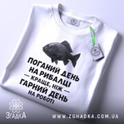 Футболка рибалка жарт. Біла футболка, складена на поверхні, зображена з верхнього ракурсу. Фон світло-сірий. На футболці чорний принт з текстом і малюнком риби, а також зелена етикетка.
