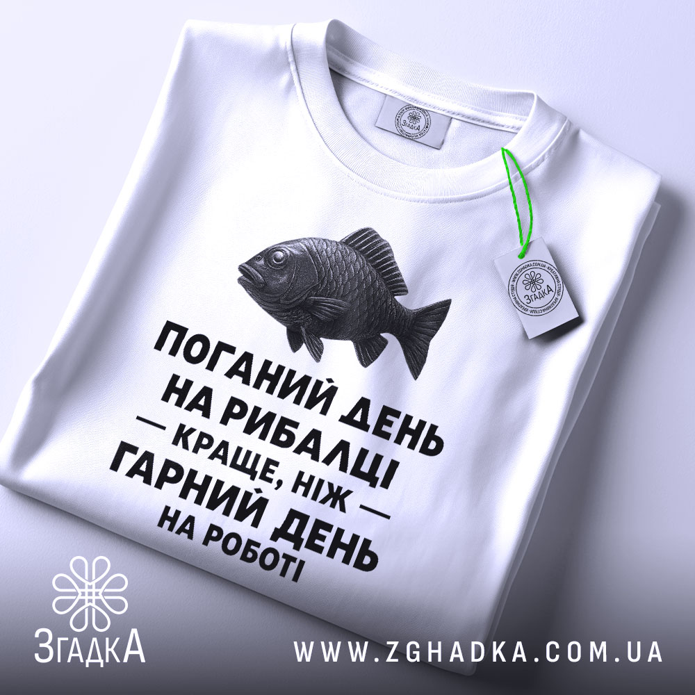 Футболка рибалка жарт. Біла футболка, складена на поверхні, зображена з верхнього ракурсу. Фон світло-сірий. На футболці чорний принт з текстом і малюнком риби, а також зелена етикетка.