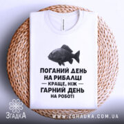 Футболка рибалка жарт. Біла футболка, складена на плетеній основі, зображена з верхнього ракурсу. Фон світло-сірий. Принт з чорним текстом і малюнком риби розташований по центру.