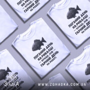 Футболка рибалка жарт. Біла футболка з коротким рукавом, зображена з близького ракурсу. На фоні видно світло-сірий градієнт. Принт з чорним текстом і малюнком риби розташований по центру.