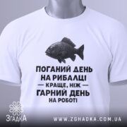 Футболка рибалка жарт. Біла футболка, складена на поверхні, зображена з верхнього ракурсу. Фон світло-сірий. На футболці чорний принт з текстом і малюнком риби, а також зелена етикетка.