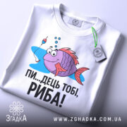 Футболка рибальський настрій. Біла футболка, складена вдвічі, з принтом риби, що має великі очі і усмішку, поряд з поплавком. Текст чорного кольору: 'ПИ...ДЕЦЬ ТОБІ, РИБА!'. На футболці є етикетка з зеленим шнурком. Фон світлий, з легким відблиском.