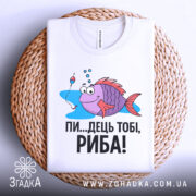 Футболка рибальський настрій. Біла футболка, складена на плетеній кошику, з принтом риби, що має усмішку і поплавок поруч. Текст чорного кольору: 'ПИ...ДЕЦЬ ТОБІ, РИБА!'. Фон світлий, що підкреслює текстуру футболки.