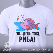 Футболка рибальський настрій. Біла футболка, складена вдвічі, з принтом риби, що має великі очі і усмішку, поряд з поплавком. Текст чорного кольору: 'ПИ...ДЕЦЬ ТОБІ, РИБА!'. На футболці є етикетка з зеленим шнурком. Фон світлий, з легким відблиском.