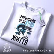 Футболка риболовля це життя. Біла футболка, що лежить на поверхні, з видимим ярликом. Ракурс зверху, фон світлий, принт з рибою та текстом яскравий.