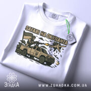 Футболка сафарі на окупанта, білого кольору, складена на столі, фон світло-сірий, принт з військовим, що стріляє з автомобіля, з українським прапором, на етикетці зелена стрічка.