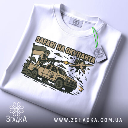 Футболка сафарі на окупанта, білого кольору, складена на столі, фон світло-сірий, принт з військовим, що стріляє з автомобіля, з українським прапором, на етикетці зелена стрічка.