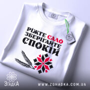 Футболка сало і спокій, біла футболка складена на поверхні, фон світло-сірий, принт з текстом "Ріжте сало зберігайте спокій" у червоному та чорному кольорах, з додатковими графічними елементами, на футболці є етикетка.