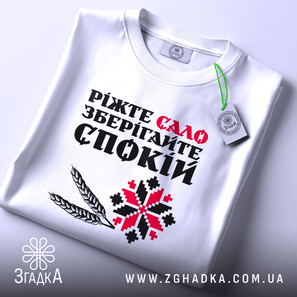 Футболка сало і спокій, біла футболка складена на поверхні, фон світло-сірий, принт з текстом "Ріжте сало зберігайте спокій" у червоному та чорному кольорах, з додатковими графічними елементами, на футболці є етикетка.