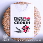 Футболка сало і спокій, біла футболка складена на плетеній кошику, фон світло-сірий, принт містить текст "Ріжте сало зберігайте спокій" з червоними та чорними літерами, а також графічні елементи у вигляді колосків і візерунка.